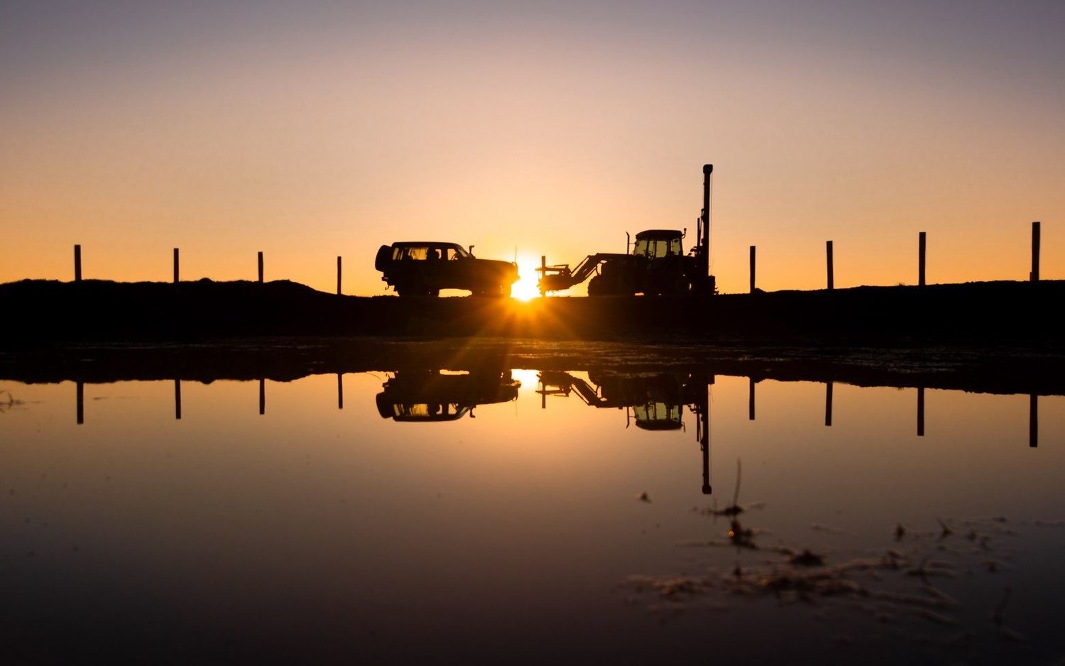A new fence line at dusk, posts to the horizon, the ute and post driver mirrored in water