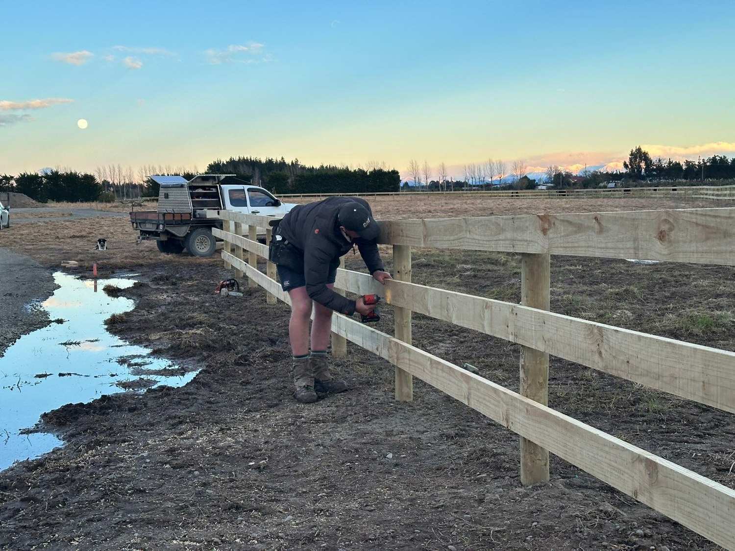 Nathan fixing a rail on a new post and rail fence, the ute and dog behind him