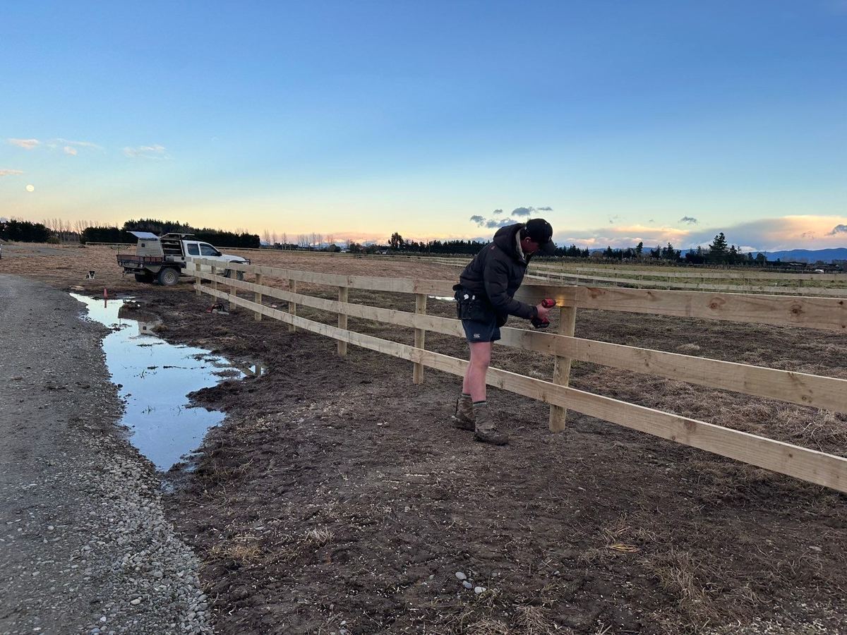 Nathan fixing a rail on a new post and rail fence, the ute and the dog behind, snow on the Alps