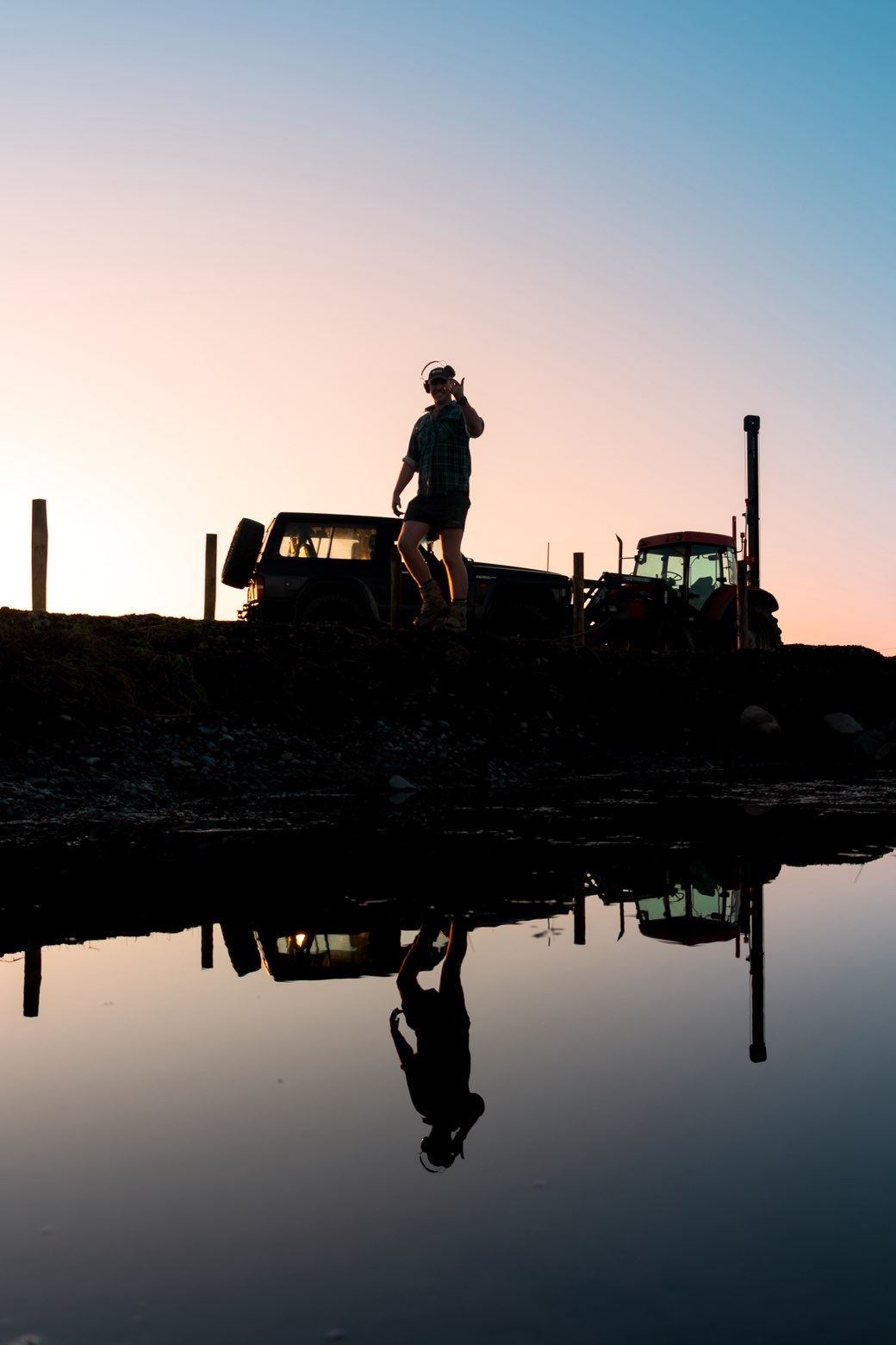 Nathan between the four-wheel drive and the tractor at dusk, reflected in still water